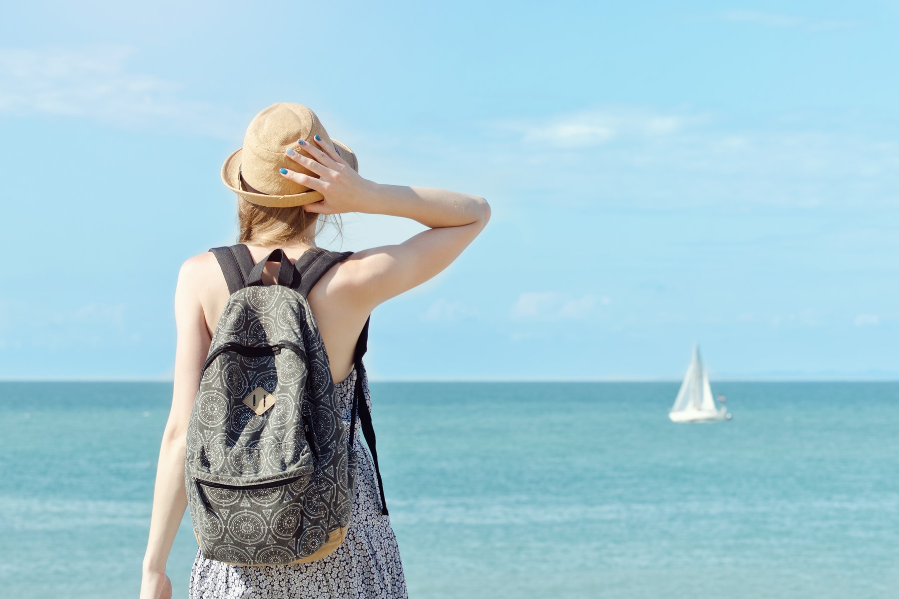 A girl in a hat with backpack standing looking out at boat on water