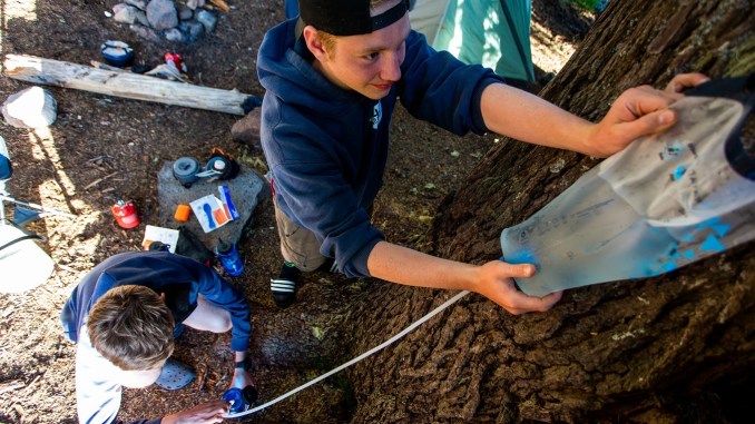 Person hangs a gravity filter at camp while another person filters into water bottle