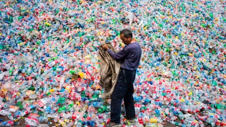 Photo of man with bag standing in pile of plastic bottles and microplastics landfill