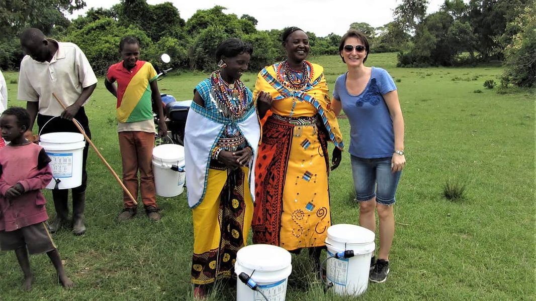 Three women stand in front of Sawyer bucket water filtration with other people using behind