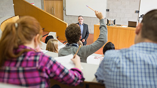 View of students in a lecture theatre. One student is raising their hand as the lecturer points to them. Representation of student engagement