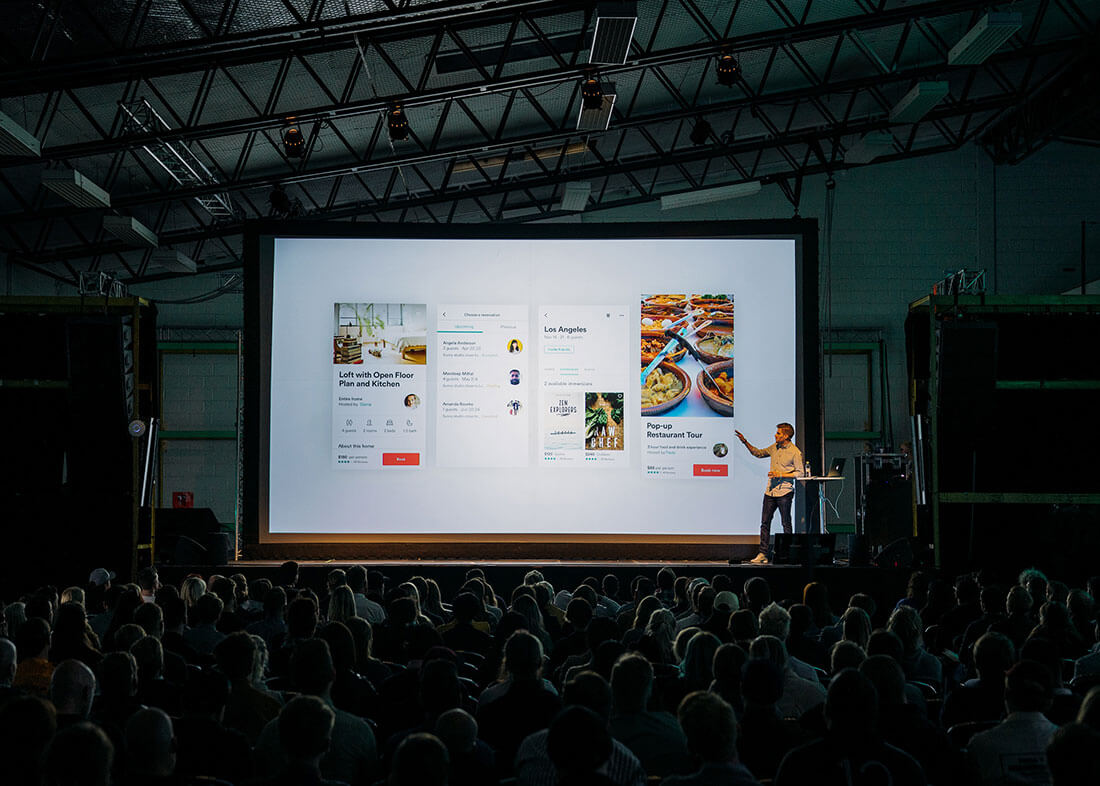 The image contains a person presenting and speaking in public in front of an audience