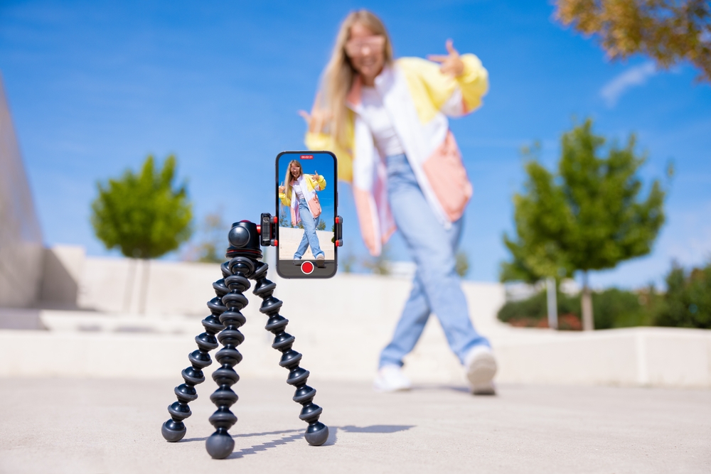 Closeup of a tripod holding a cell phone while a girl with blonde hair dances in the background.