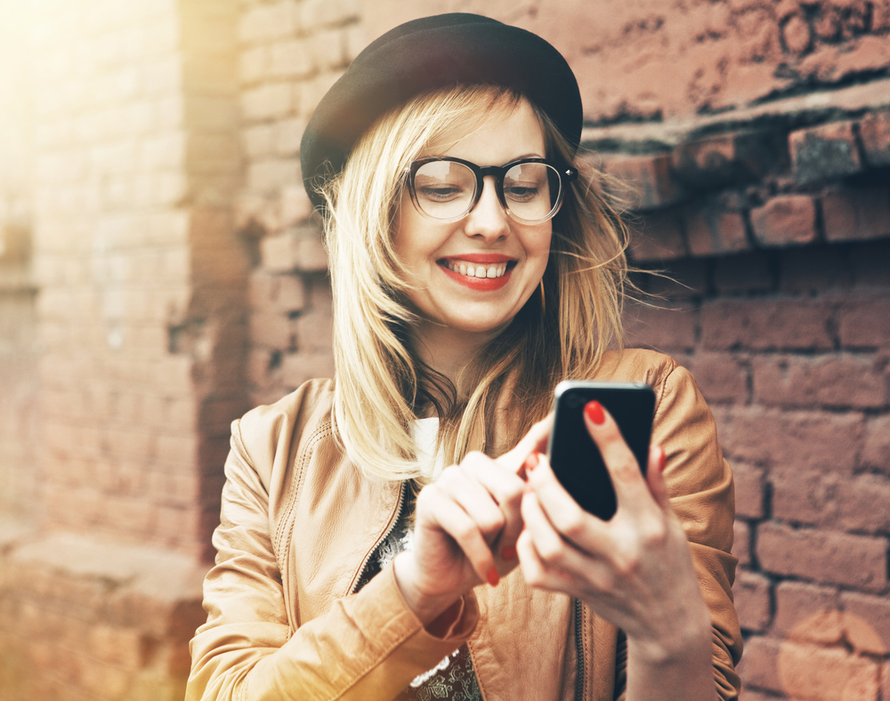 Young blonde woman wearing a bowler hat and glasses on her phone.