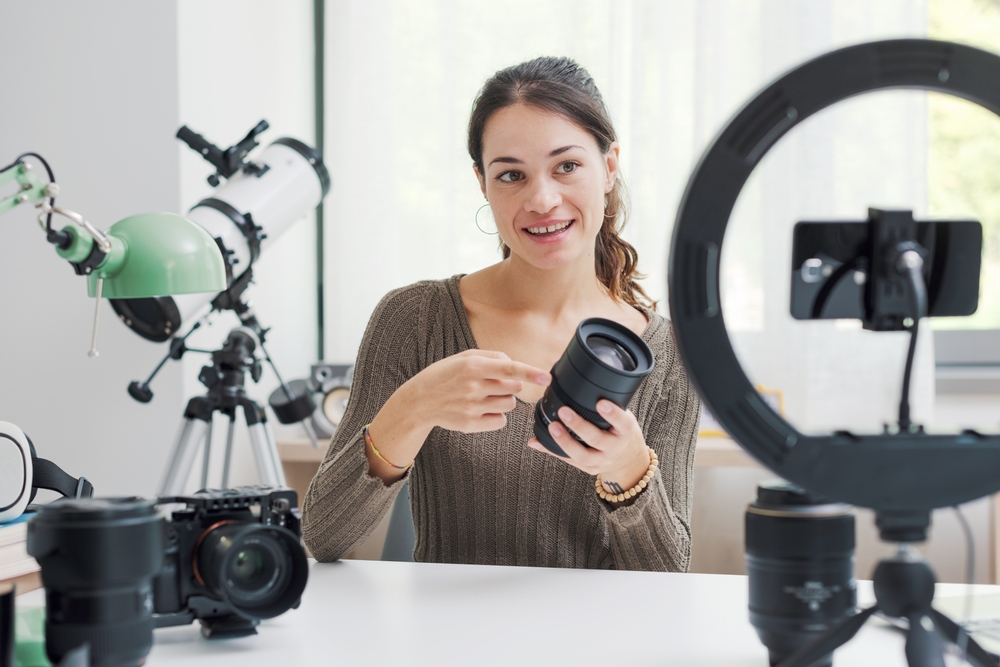 Brunette woman holding a camera lens with other camera parts on a table in front of her while a phone on a tripod records her.