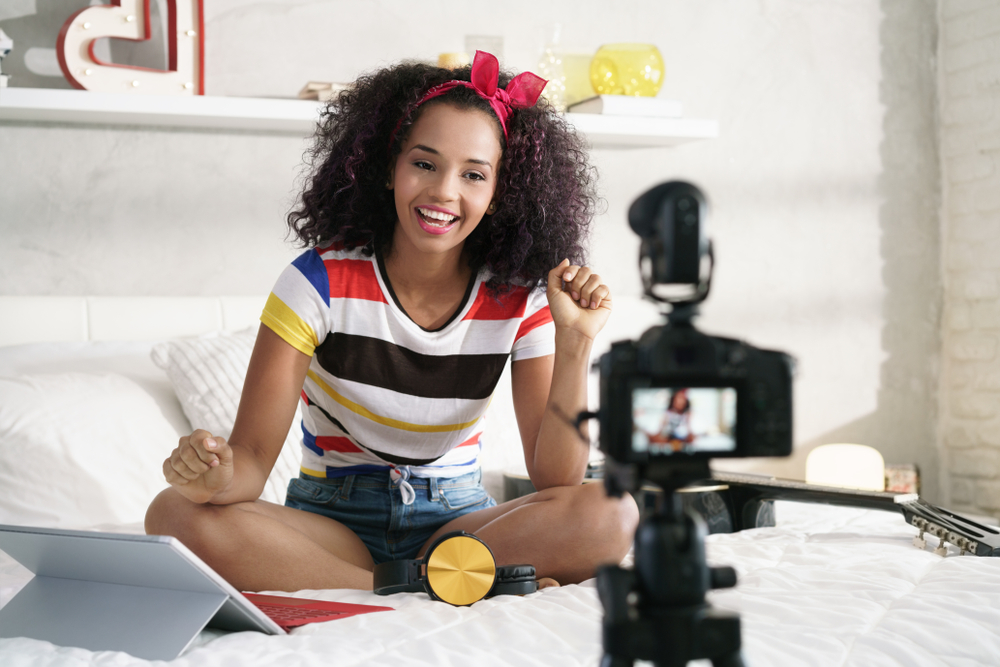 Black girl with a colorful top sitting on her white bed recording herself with a tripod. She has her laptop open next to her.