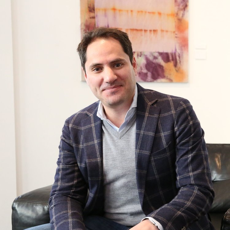 Photo of a man, Paul Strachman, in a navy checkered coat and grey sweater sitting on a black leather couch