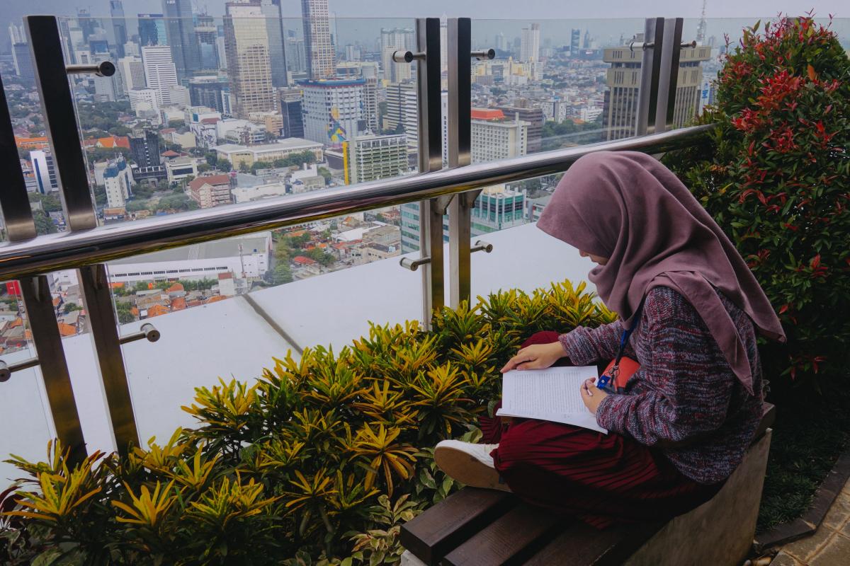 Student wearing a hijab sits near a window and reads. Cityscape in the distance.