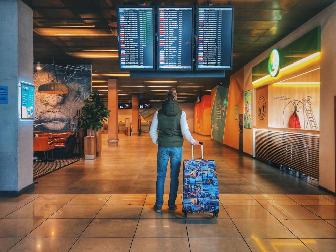 foreign exchange student holds suitcase and looks up at an airport flight information screen.