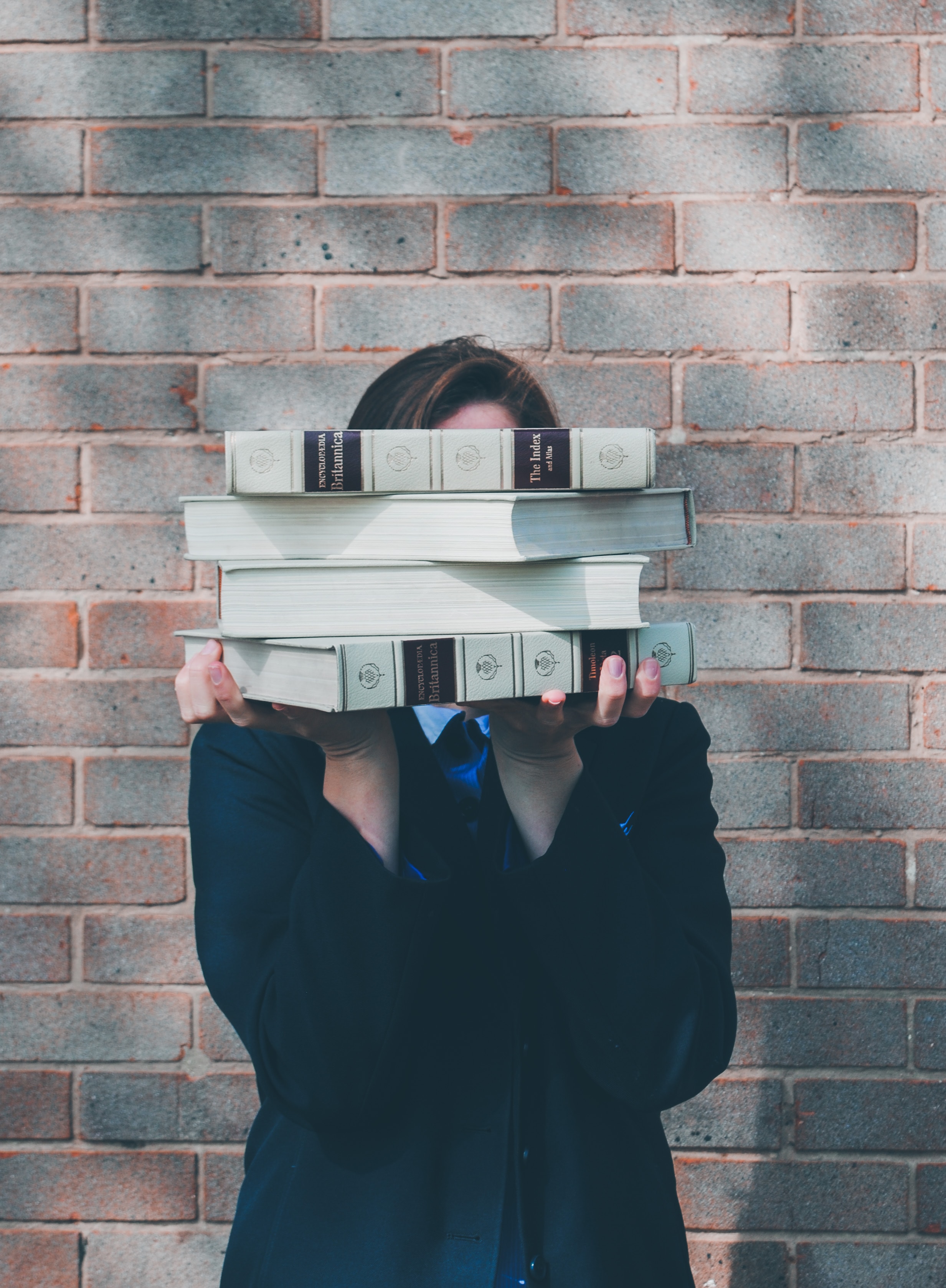 A high school student holds a stack of Encyclopedias in front of their face, while standing in front of a brick wall. 