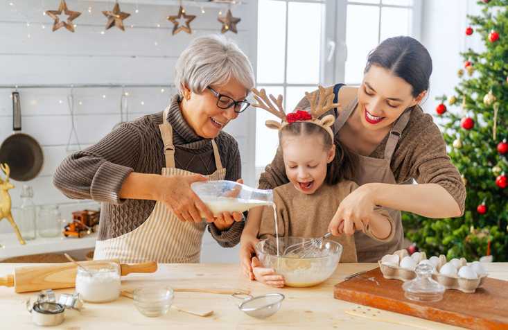 Three generations of people baking cookies together in a kitchen with a Christmas tree in the background