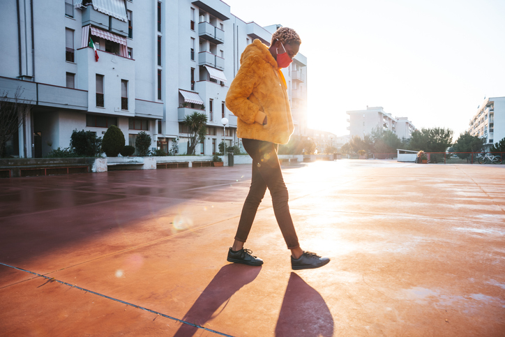 Person in an orange, fuzzy jacket and wearing a face mask walking on an empty walkway in front of a residential building. The morning sun is peaking in the background.