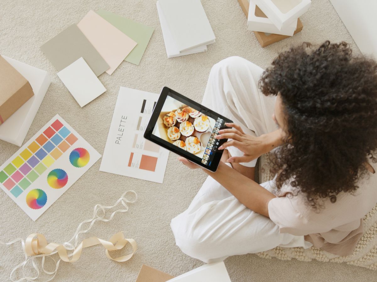 Overhead shot of a woman sitting on the floor, surrounded by colour swatches, sheets of card stock paper and boxes, while she edits an image of a box of cupcakes on her iPad.