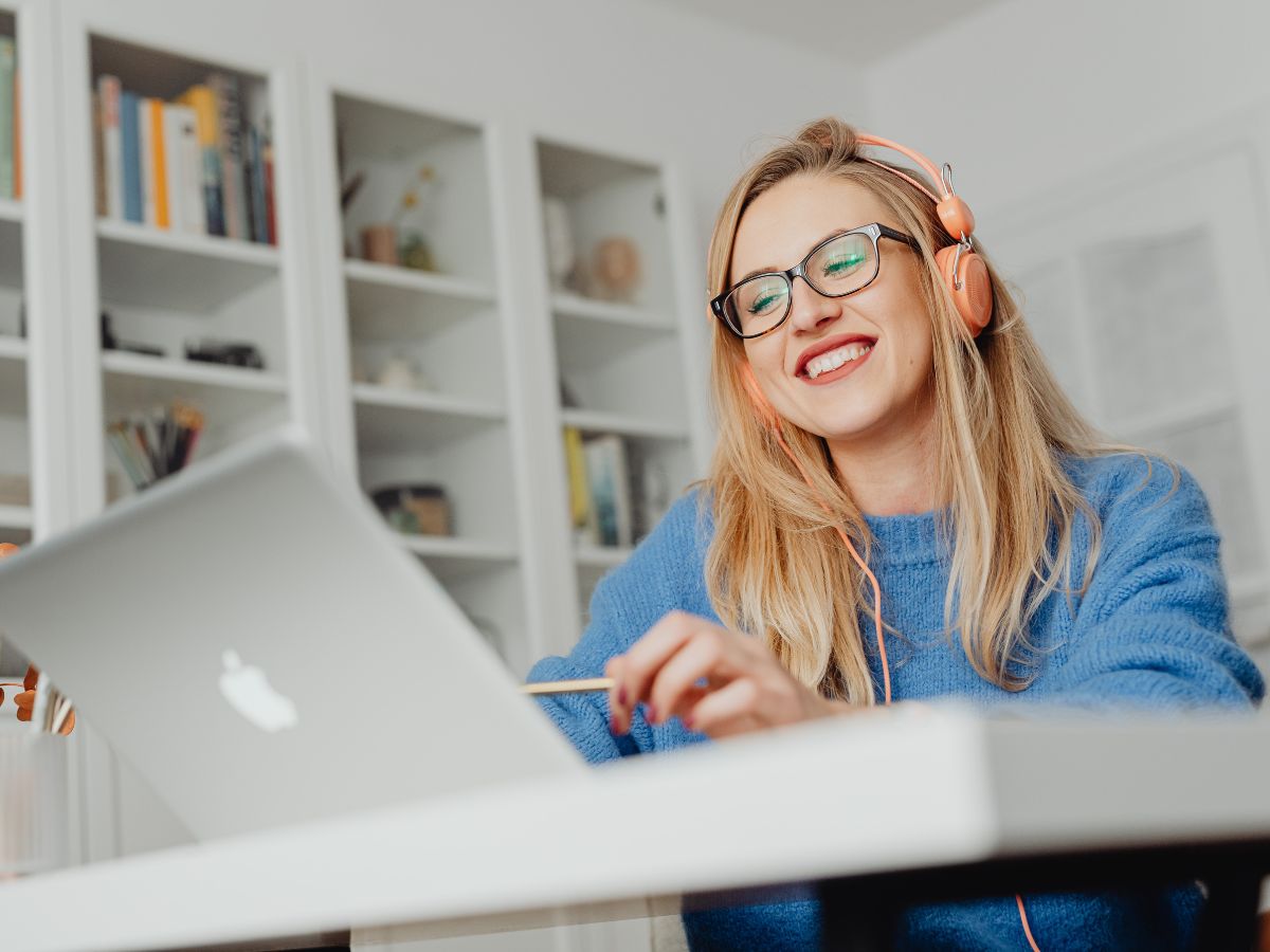 A smiling woman wearing glasses and a blue jumper, listening to something on her pink headphone while looking at something in her silver laptop.
