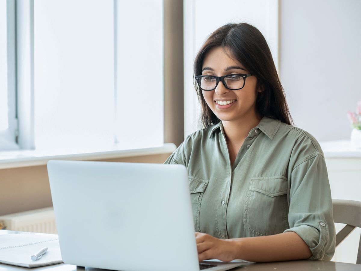 A smiling woman wearing glasses and an olive green button down shirt as she looks at something in a silver laptop. 