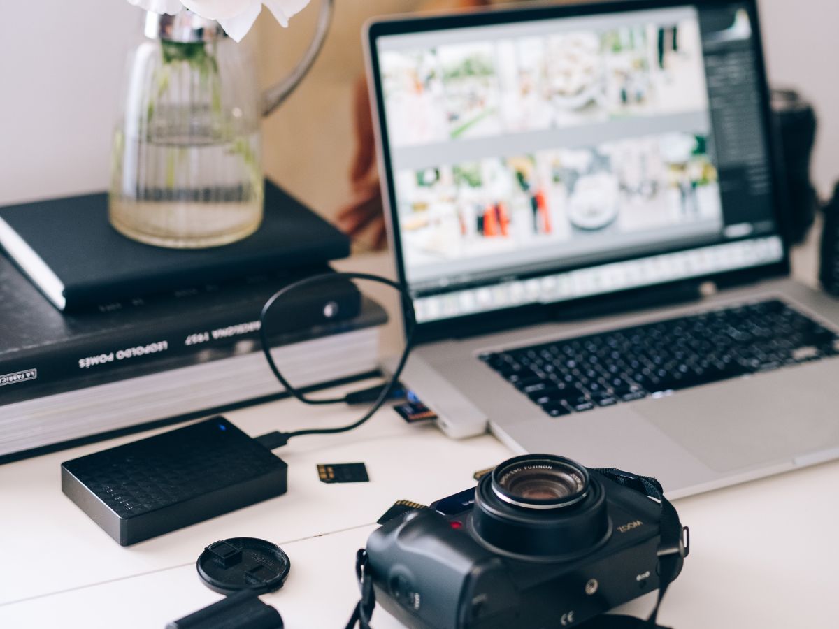 A silver laptop with a hard drive connected to it as it uploads photos. Next to the hard drive is a black camera with the lens cover off. All objects are in a white surface.