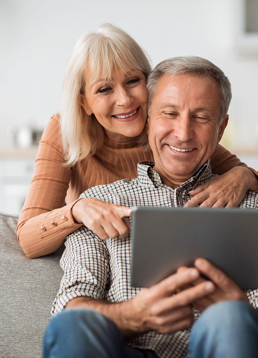 older woman hugging older male over the shoulder while he looks at a tablet