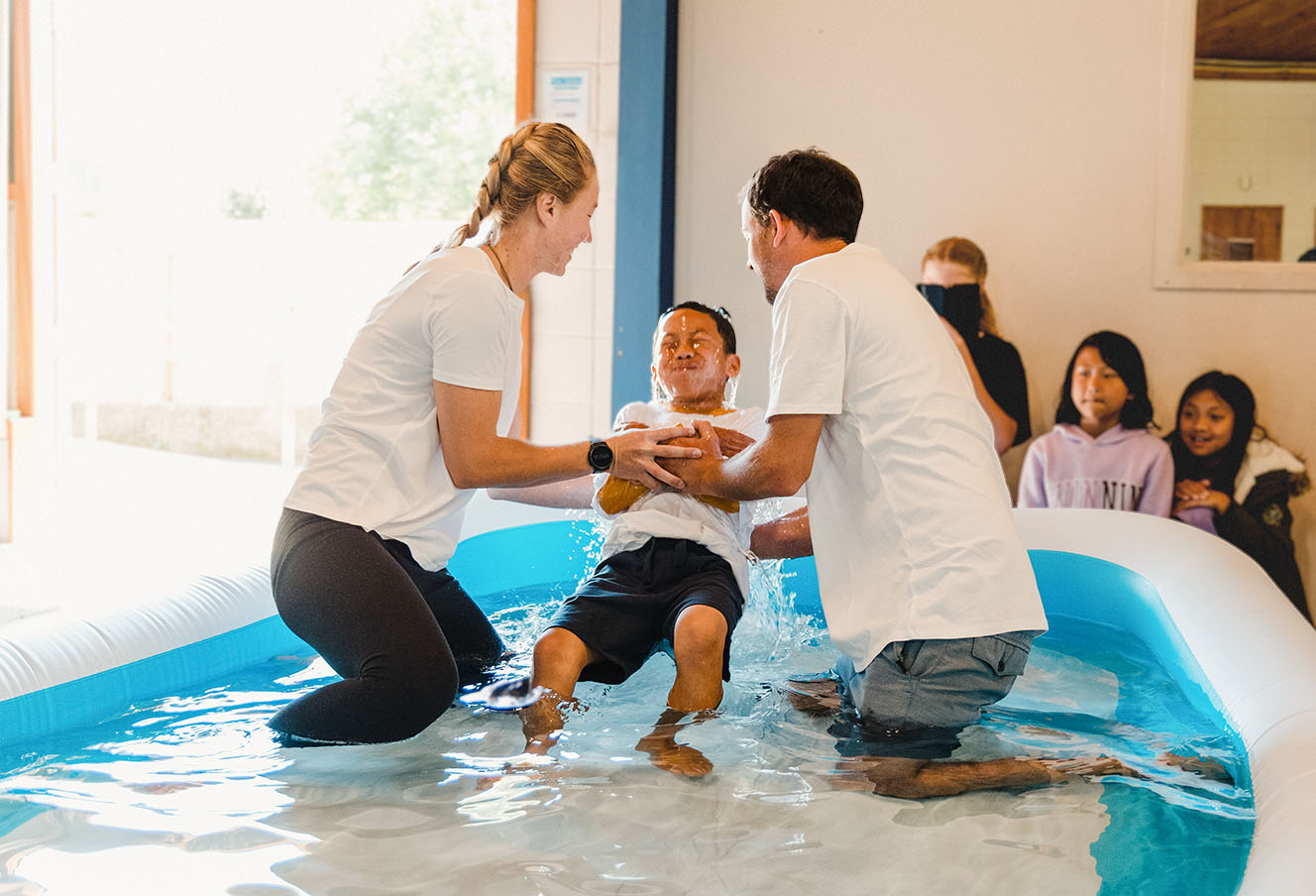 two young adults baptise a smiling child in an inflatable pool in a church with other children looking on