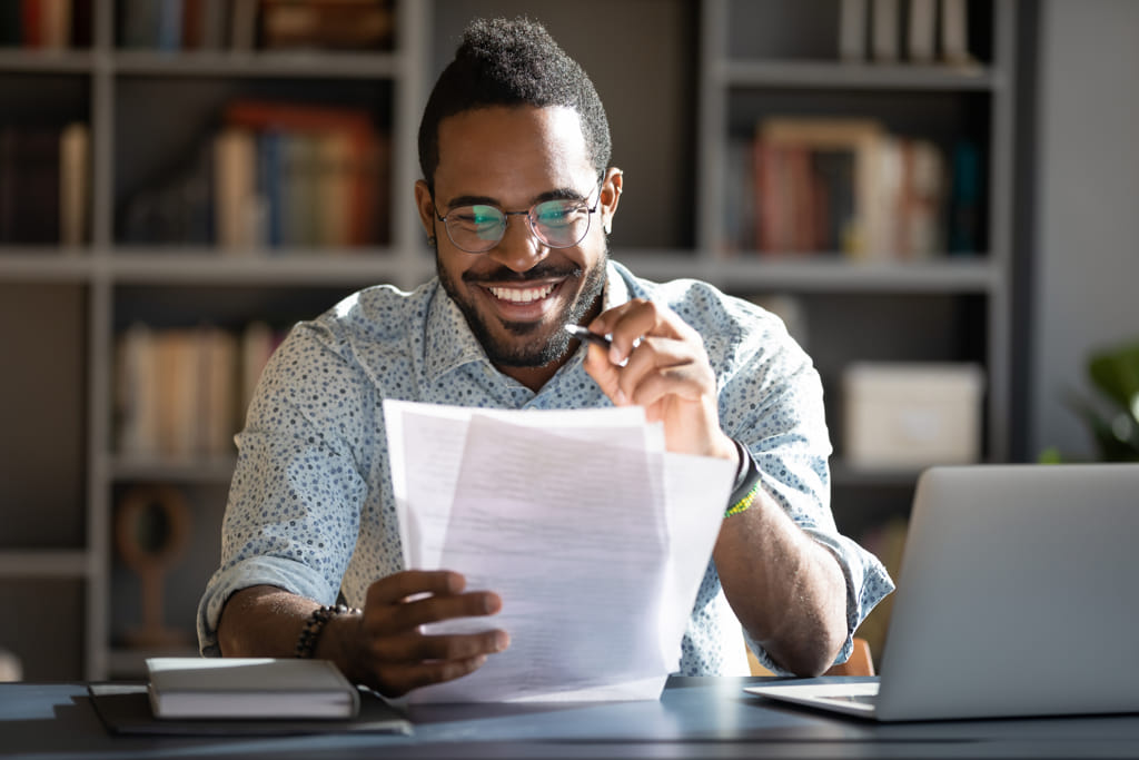 African student guy sitting at desk holding papers printed tasks perform test prepares for entrance exams enjoy process of study