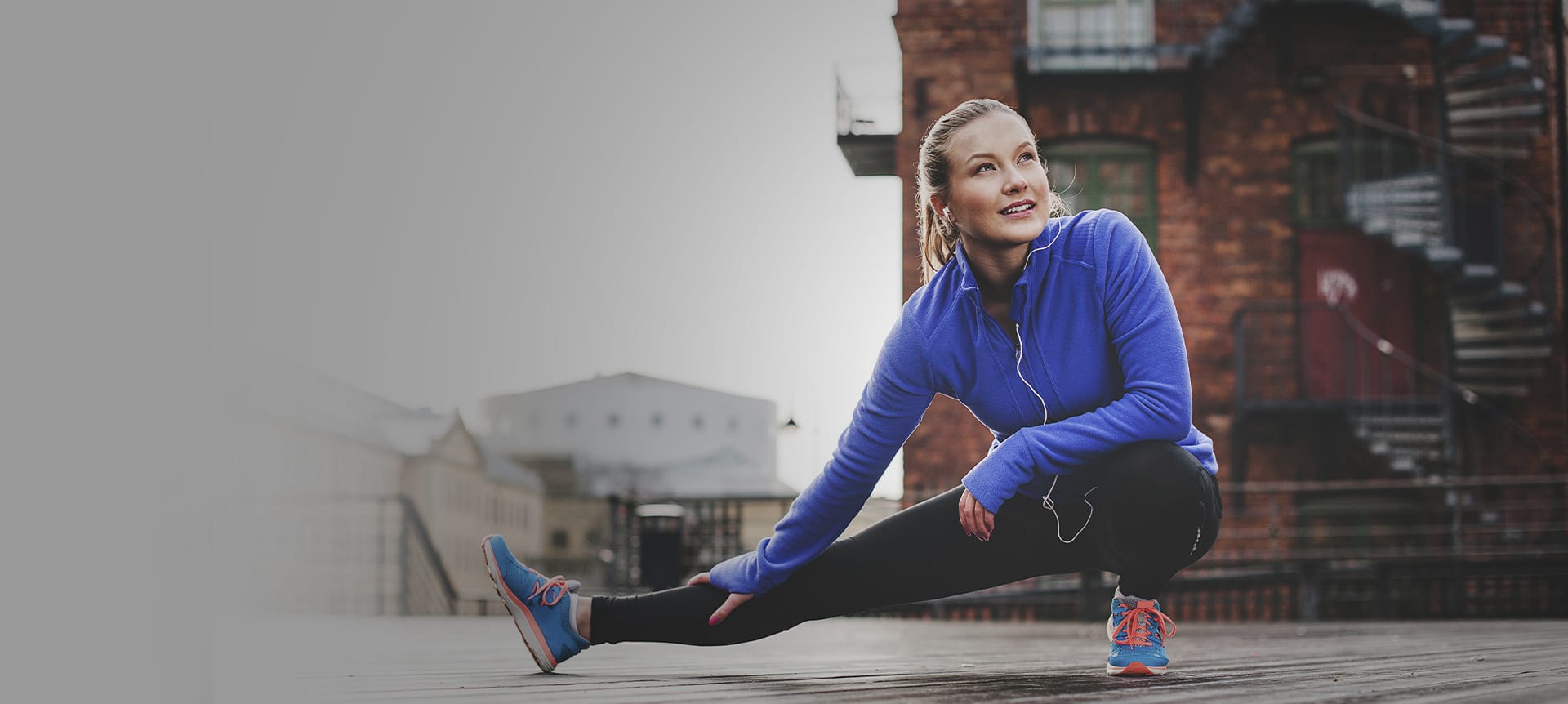 Eine Frau in einer blauen Trainingsjacke dehnt ihr rechtes Bein mit einem Ausfallschritt vor einem Backsteingebäude.