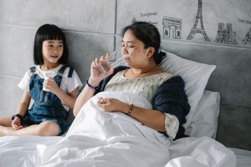 A child caregiver archetype sits with an elder who is in bed, drinking a glass of water.