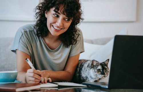 Smiling person writes in a notebook. A cat sits next to them.