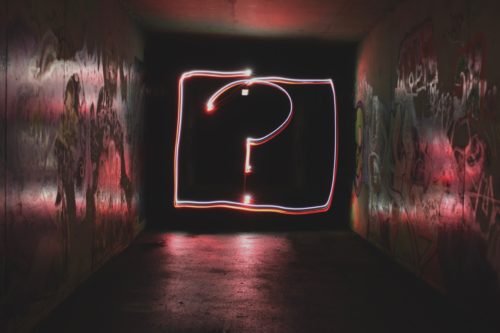 A neon question mark inside a neon square against a black background at the end of a metallic hallway.