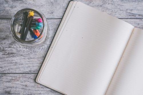 A blank journal and a jar of multi-colored pens on a gray wood surface.