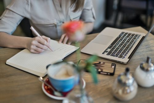 A person writing in a notebook at a desk beside a laptop computer and a blue teacup.