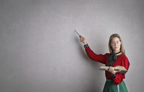 Woman wearing glasses holding book and pointing to a blank chalkboard.