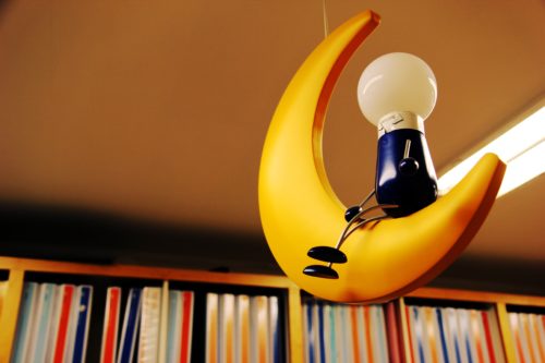 A light fixture in the shape of a person sitting on the moon hangs in front of a bookcase.