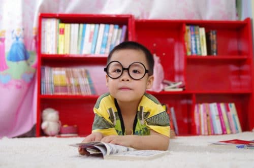 Child with big glasses sits at a table in front of a full bookshelf, reading a children's book.