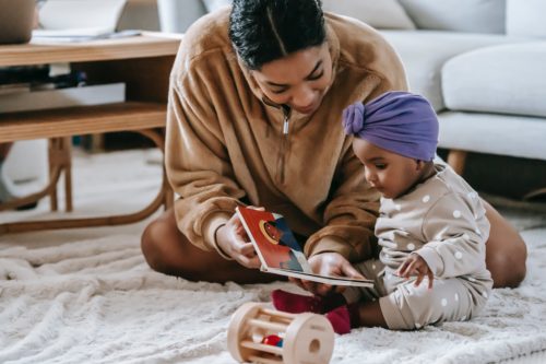 A woman sits on the floor beside a baby, reading a picture book to the child.