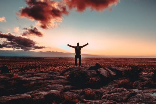 A person standing in a desert landscape at sunset with arms outstretched.