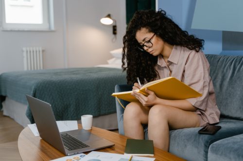 Person sitting on a blue couch writing in a large yellow journal in front of a coffee table with a laptop and journals on it.