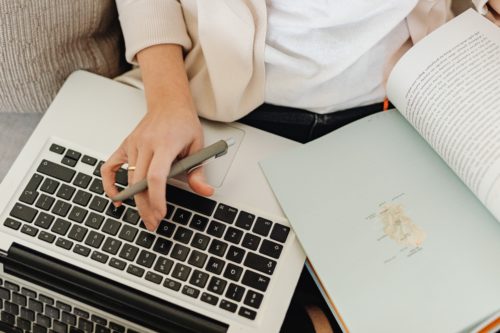 A writer holds a pen and rests one hand on a computer while flipping through a reference book with the other.