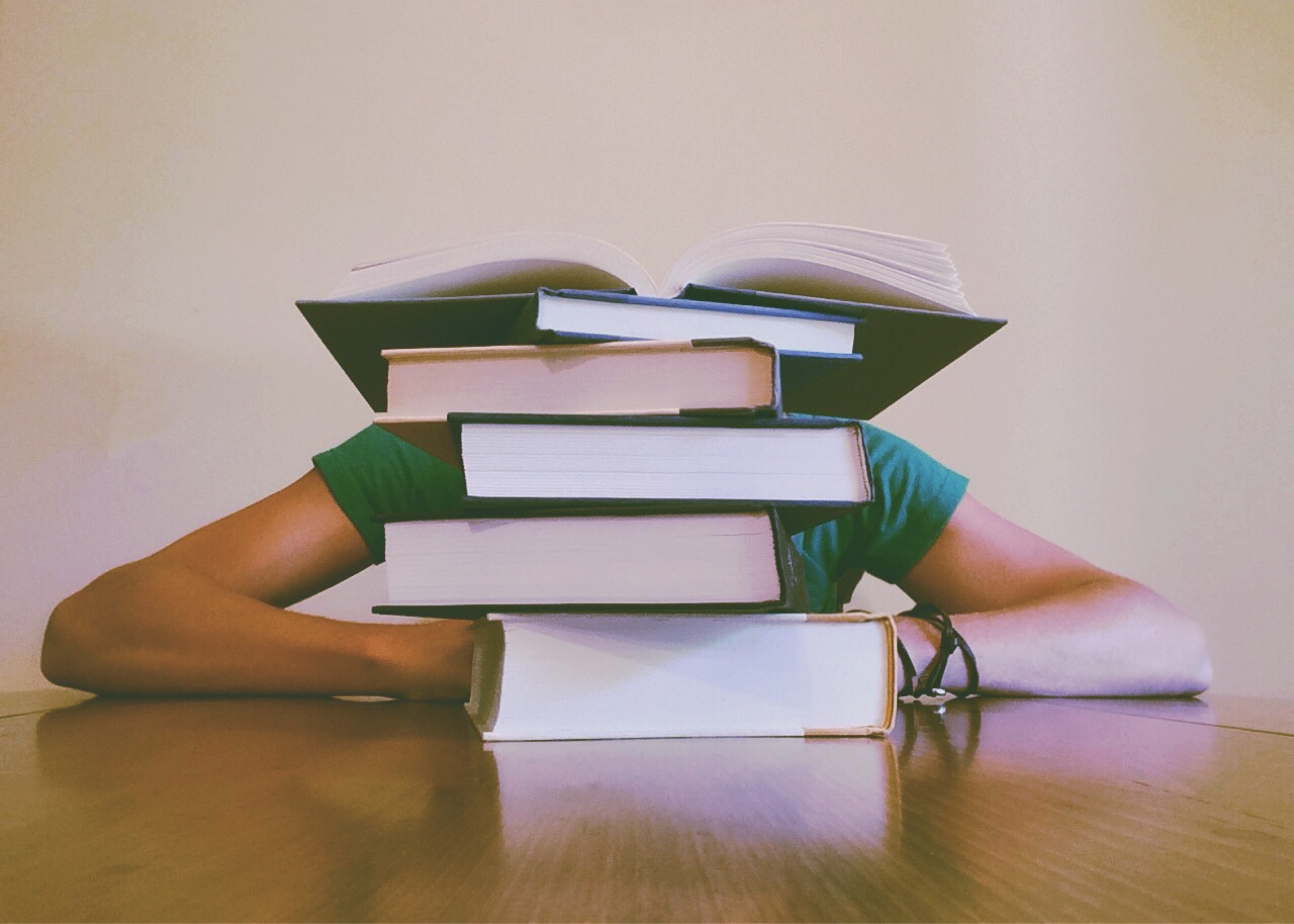 A person in a green shirt at a table, their face hidden behind a stack of books.