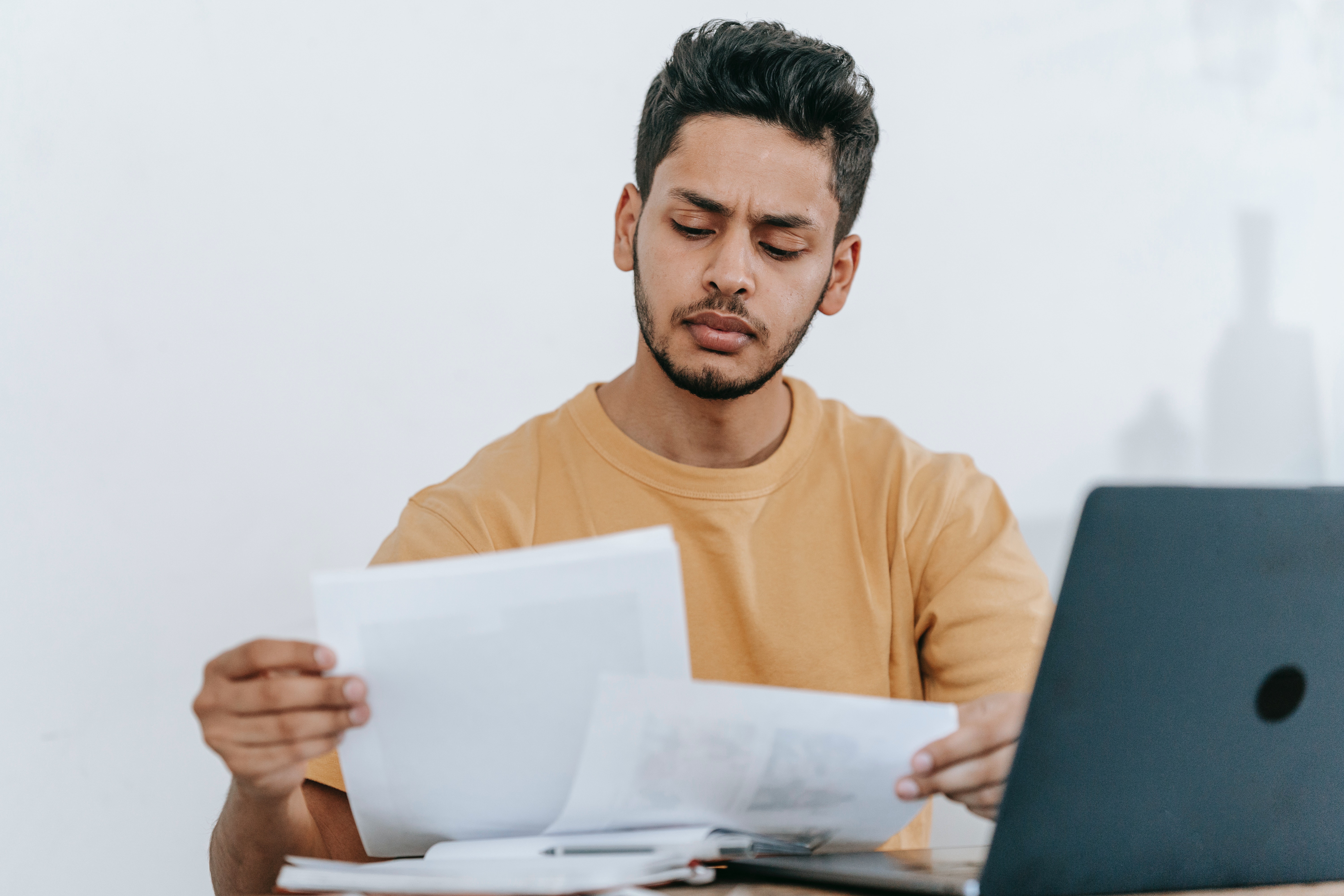 A person in a yellow shirt sits in front of a laptop, frowning at a stack of papers they're holding.