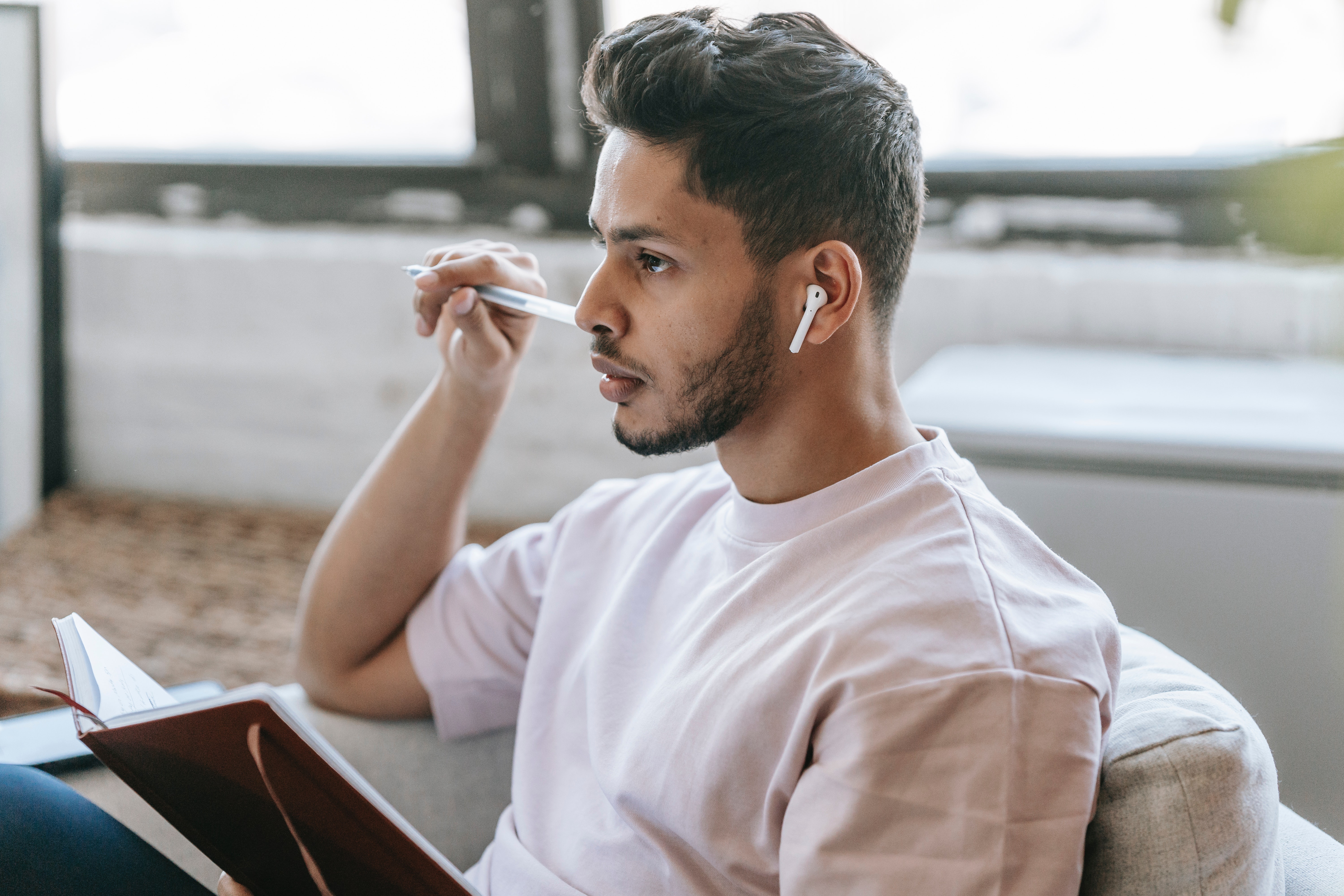A person with a beard and wearing earbuds holds a leather journal and rests a pen against their cheek as they stare off into the distance.