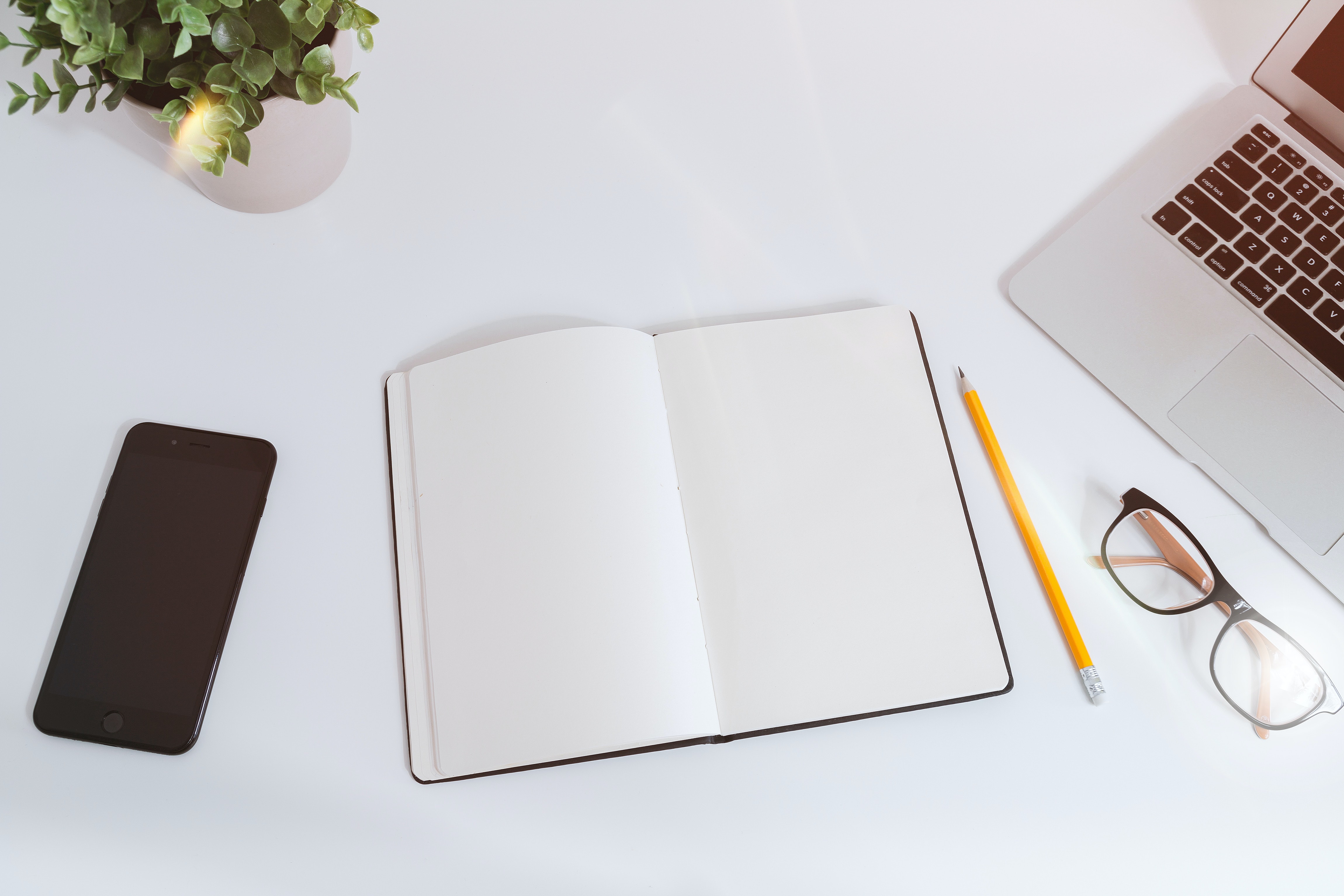 An overhead view of a blank notebook, black phone, yellow pencil, glasses, laptop, and houseplant on a white desk..