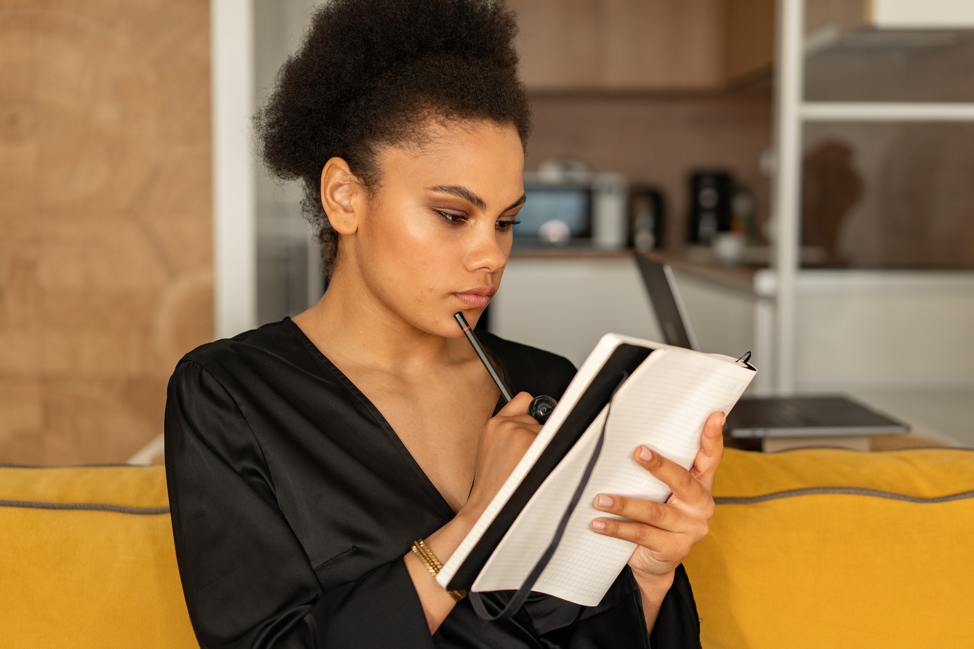 A female-presenting person sits on a yellow couch and touches a pencil to their chin and thinks as they look at a notebook.