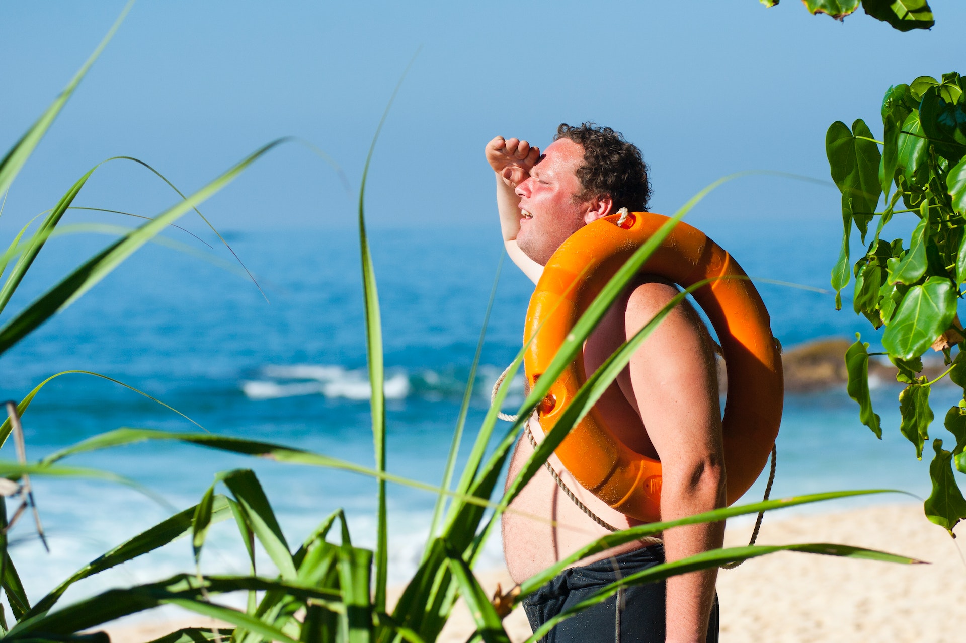 A person with life preserver looped around their arm stands on a beach wearing swim trunks and shielding their eyes with their hand as they search for something in the distance.