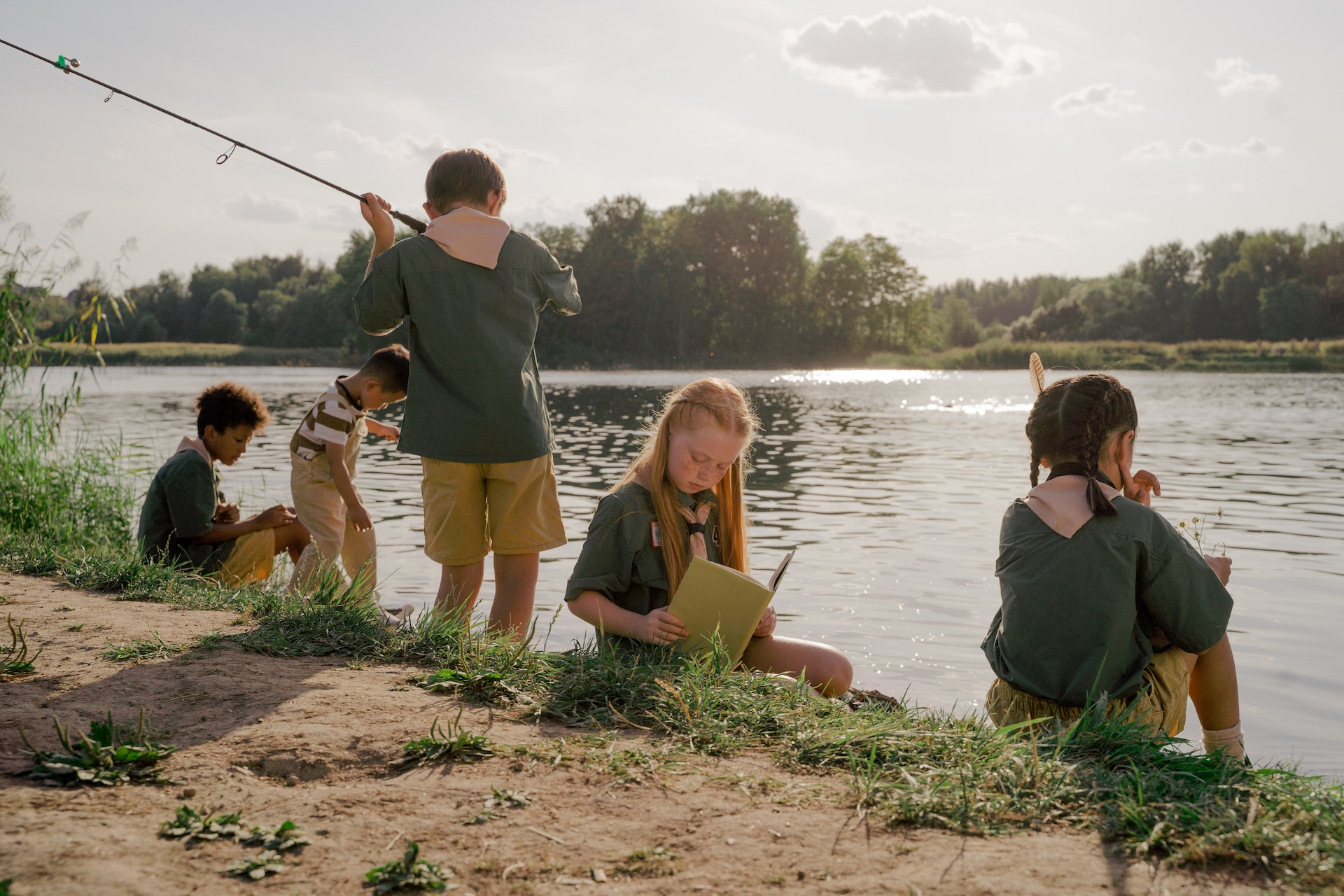 Kids sit along the shore of a lake, reading and fishing.