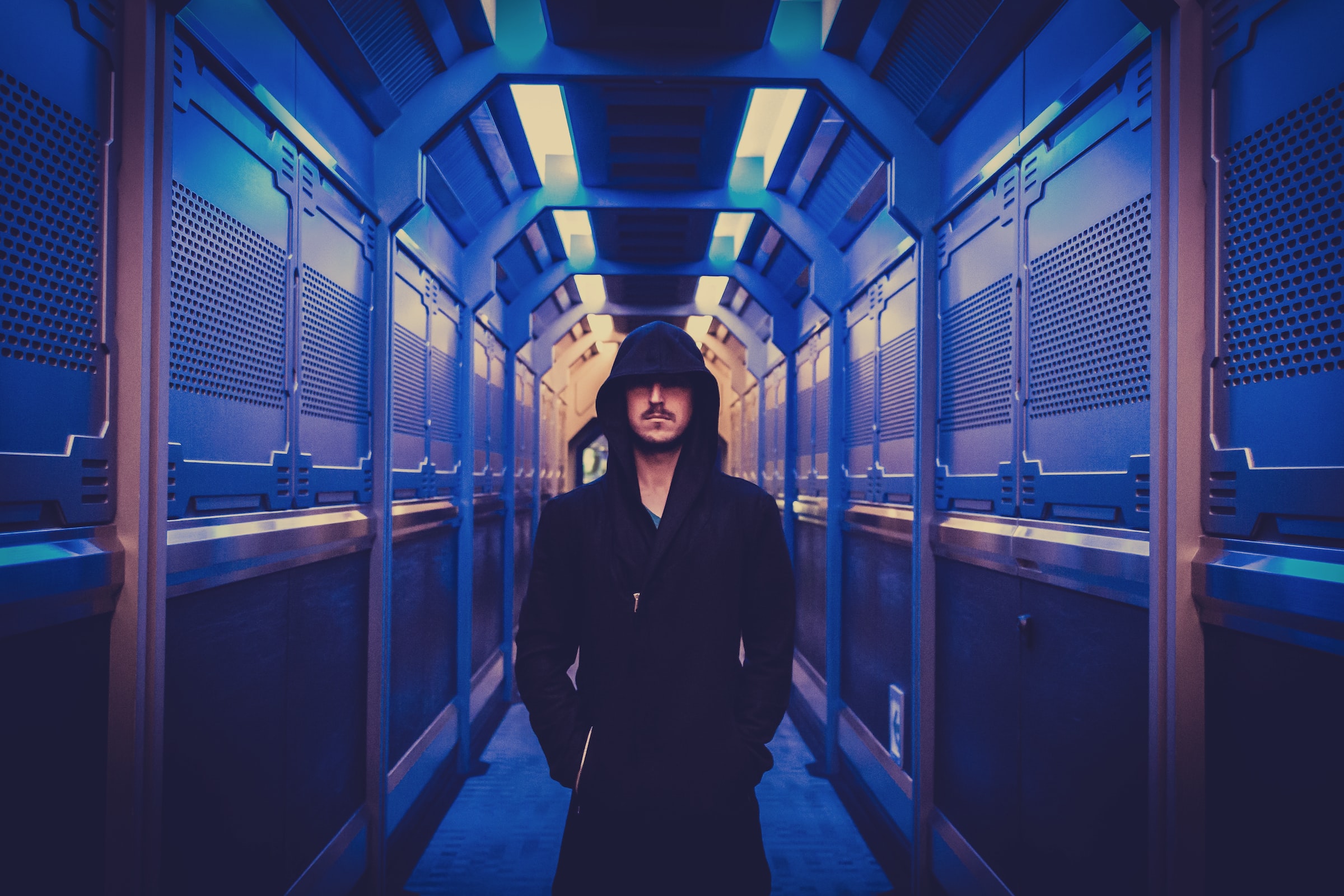 A person in a black hoodie walks down a blue metallic hallway.