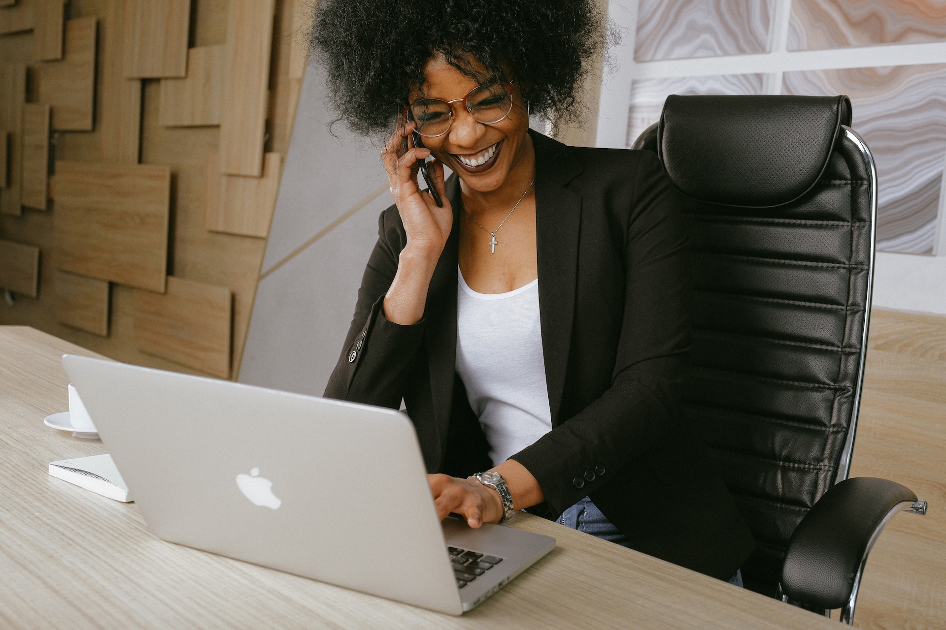 A smiling agent in business attire sits at a desk talking on the phone and looking at a computer screen.
