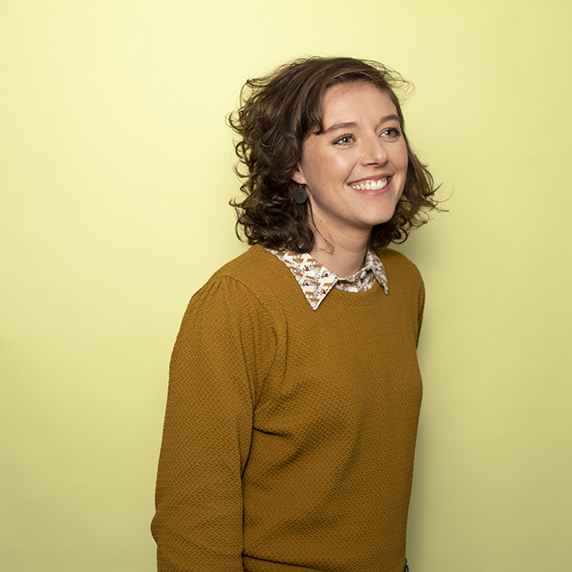 Vrouwelijke IT Trainee kijkt lachend in de camera bij een gele achtergrond