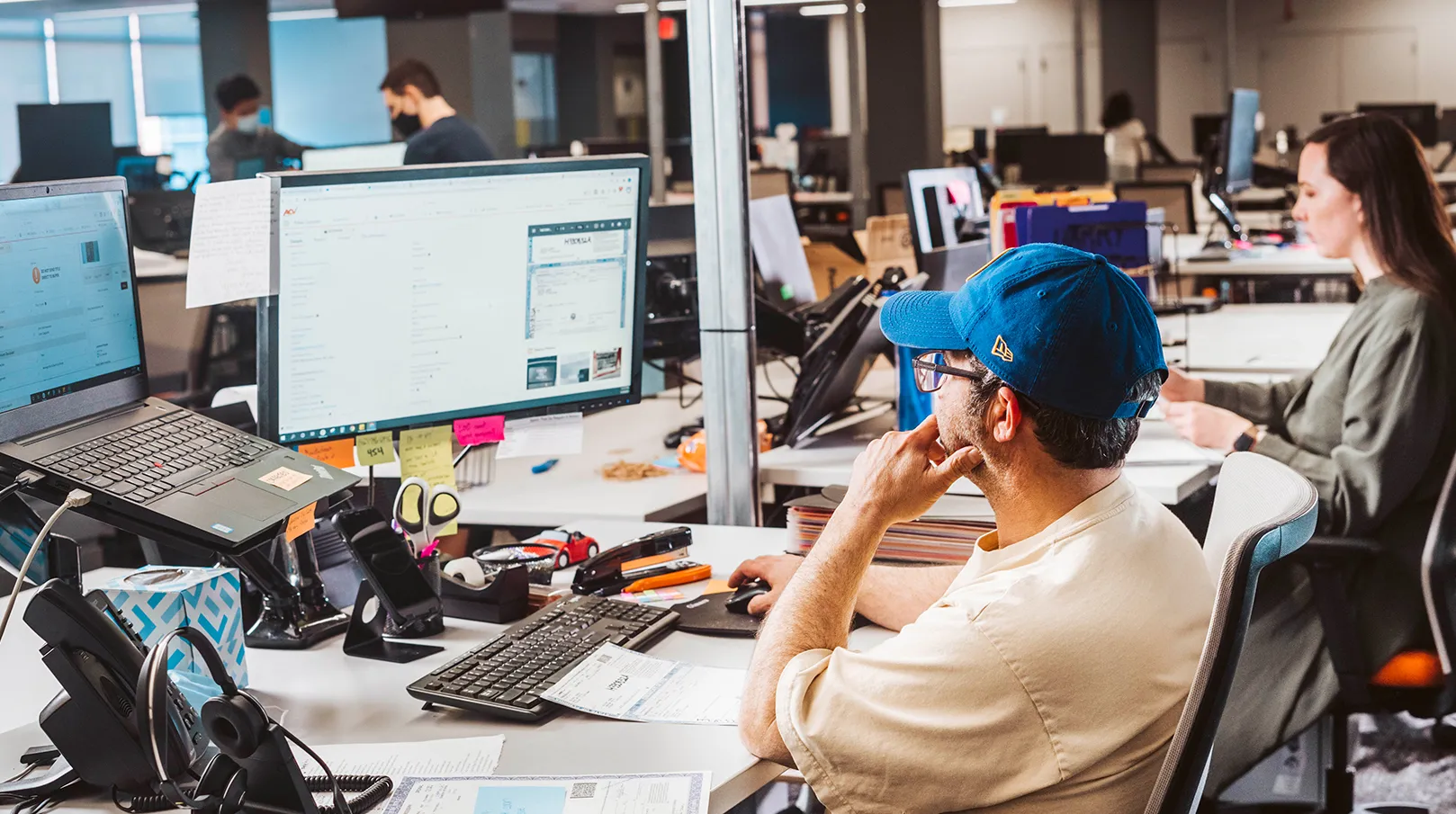 A man wearing a blue hat and yellow shirt sits at his desk looking at his computer monitor in a populated office.