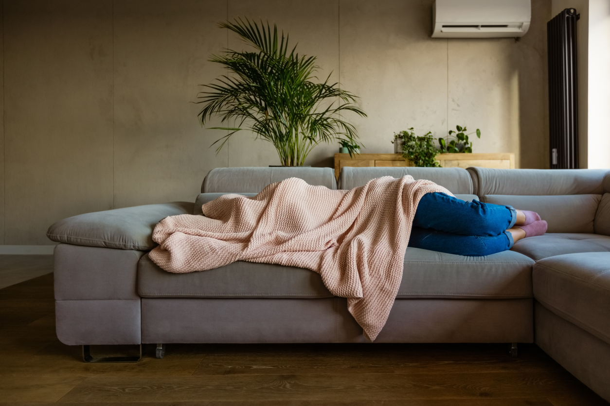 Person laying on a couch covered with a blanket with sun shining through a window