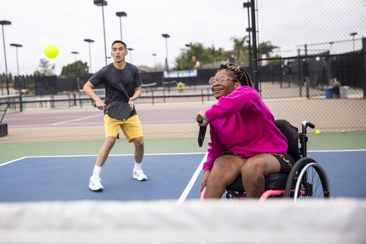 A young black woman in a wheelchair playing pickleball with her friend.