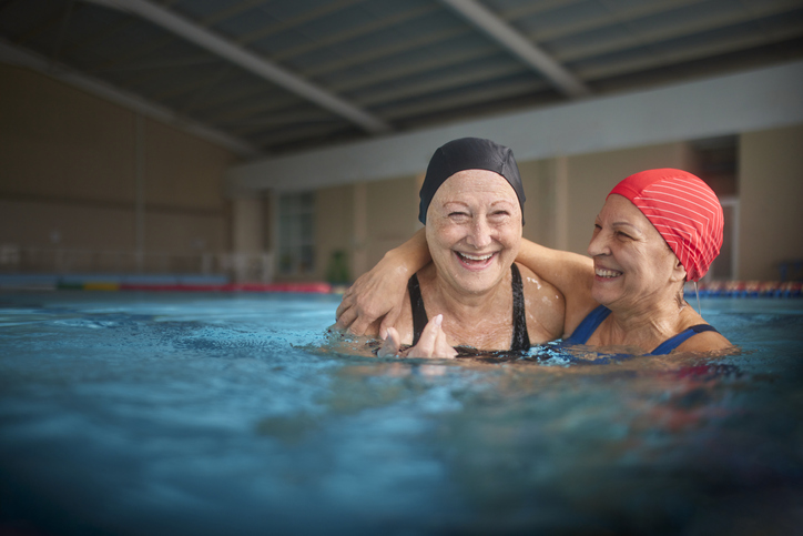 Two mature women laughing in a pool, wearing bathing suits and caps